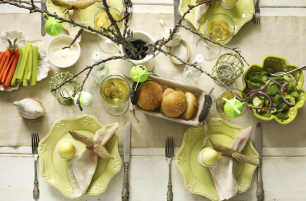 A green photo of an overhead view of a white table set for easter with pastel plates and bunnies. Branches with easter eggs dangling from them fill a vase at the centre of the table. 